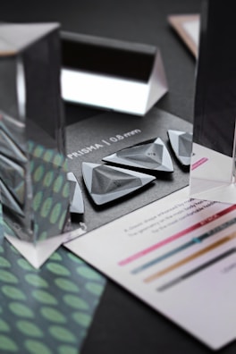 An arrangement of triangular and rectangular prisms sits on a dark background. The prisms are transparent and reflective, creating visually interesting reflections and refractions. Surrounding the prisms are pieces of paper with text and colored lines. A pattern of green and teal dots is visible on one sheet.