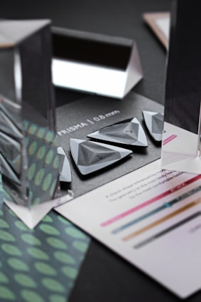 An arrangement of triangular and rectangular prisms sits on a dark background. The prisms are transparent and reflective, creating visually interesting reflections and refractions. Surrounding the prisms are pieces of paper with text and colored lines. A pattern of green and teal dots is visible on one sheet.