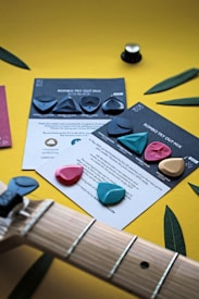 A collection of guitar picks of various shapes and colors are displayed on promotional cards against a vibrant yellow background. The picks include shades of blue, pink, and black. Part of a guitar neck is visible in the foreground. Green leaves add a natural touch to the composition.