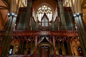 A large and ornate pipe organ dominates the interior of a church, featuring intricate wooden carvings and numerous vertical pipes. The vaulted ceiling and stained glass windows add to the grandeur, with gothic architectural elements present throughout.