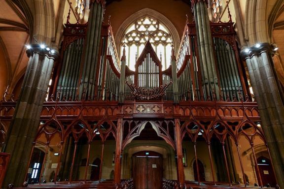 A large and ornate pipe organ dominates the interior of a church, featuring intricate wooden carvings and numerous vertical pipes. The vaulted ceiling and stained glass windows add to the grandeur, with gothic architectural elements present throughout.