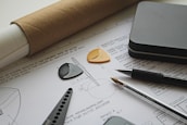 The image depicts an architectural or engineering workspace featuring technical drawings with precise measurements and annotations. Several drafting tools, including a pen, pencil, protractor, and triangular scale, are placed on top of the drawings. A rolled-up sheet of paper and a metallic rectangular case are also visible, adding to the professional and organized setting.