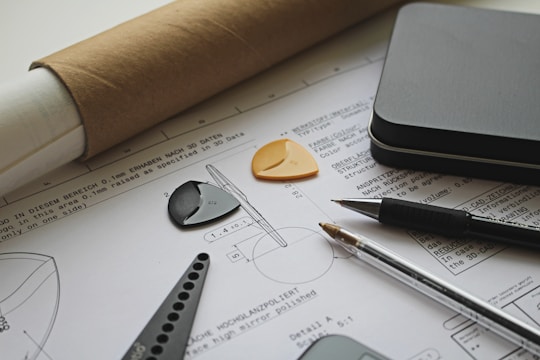 The image depicts an architectural or engineering workspace featuring technical drawings with precise measurements and annotations. Several drafting tools, including a pen, pencil, protractor, and triangular scale, are placed on top of the drawings. A rolled-up sheet of paper and a metallic rectangular case are also visible, adding to the professional and organized setting.