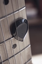 Close-up view of a guitar with a dark guitar pick wedged between the strings against the fretboard. The strings are metallic and the fretboard appears to be made of wood with visible frets. The background is blurred, focusing attention on the strings and pick.