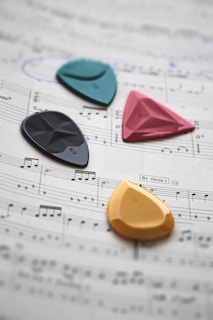 Four colorful guitar picks are placed on a sheet of music. The picks are in turquoise, pink, black, and orange, and they rest on musical notes and staff lines printed on a paper.
