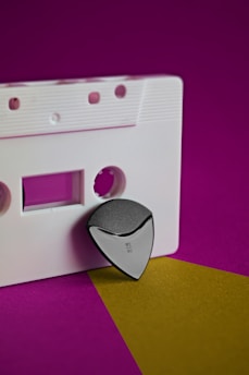 A white audio cassette appears on a vibrant purple background. Positioned at the edge of the cassette is a triangular black and silver object, giving a modern and sleek contrast to the retro feel of the cassette.