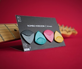 A set of four colorful guitar picks is displayed on a standing card against a rich red background. The picks are black, teal, pink, and yellow, arranged in a row. Beside the card is the neck of a guitar, partially visible. The card contains the text 'ROMBO HORIZON | 1.4 mm' along with a brand logo and a small graphic illustration.
