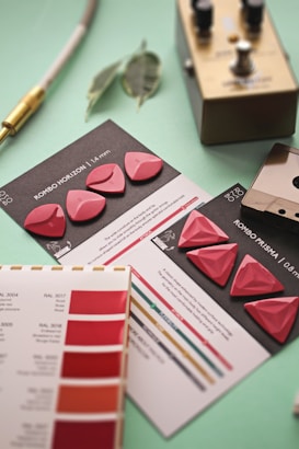 A collection of guitar picks with two distinct shapes is displayed on informational cards on a mint green surface. Nearby is a metallic effects pedal featuring knobs and switches. There are color sample cards with various shades of red and a couple of eucalyptus leaves beside a connected audio cable with a gold connector.
