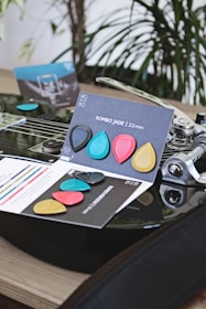 A collection of colorful guitar picks displayed on top of an electric guitar. The picks are in different colors including blue, red, yellow, and black, and are placed on a card labeled 'ROMBO JADE 2.3 mm'. The guitar and picks are set against a blurred background of green plants, creating a vibrant and artistic scene.