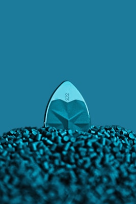 A teal-colored iron partially buried in a mound of small, cylindrical plastic pellets. The steam holes and geometric design on the iron's soleplate are visible, and the background is a solid teal color matching the iron and pellets.