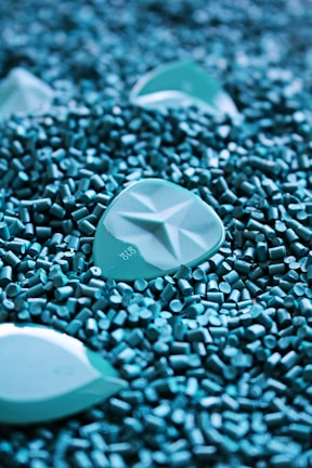 A close-up of turquoise plastic pellets with larger, similarly colored objects scattered among them. The pellets have a cylindrical shape, and the larger objects have a smooth, geometric pattern.