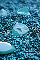 A close-up of turquoise plastic pellets with larger, similarly colored objects scattered among them. The pellets have a cylindrical shape, and the larger objects have a smooth, geometric pattern.