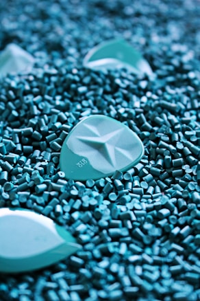 A close-up of turquoise plastic pellets with larger, similarly colored objects scattered among them. The pellets have a cylindrical shape, and the larger objects have a smooth, geometric pattern.