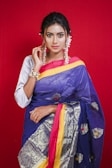 A woman poses against a bright red background, wearing a vibrant blue and gold saree with intricate patterns. Her hair is styled neatly, and she is adorned with jewelry including earrings and a bracelet. A confident and serene expression is evident on her face.