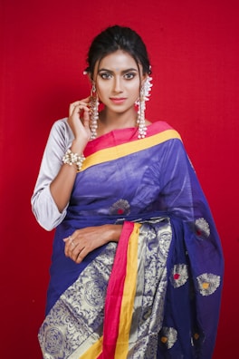 A woman poses against a bright red background, wearing a vibrant blue and gold saree with intricate patterns. Her hair is styled neatly, and she is adorned with jewelry including earrings and a bracelet. A confident and serene expression is evident on her face.