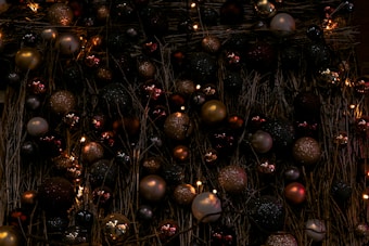 A collection of Christmas ornaments in various colors including gold, red, green, and silver are displayed together on a backdrop of intertwined dried branches. The ornaments are primarily spherical and have a glittery appearance. Some small lights are interspersed among the ornaments, creating a festive atmosphere.