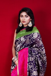 A woman stands against a plain red background, wearing a vibrant saree with intricate patterns, adorned with statement earrings and multiple bangles. The saree is primarily dark blue with metallic details, complemented by a pink border and a green pallu.