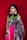A woman stands against a plain red background, wearing a vibrant saree with intricate patterns, adorned with statement earrings and multiple bangles. The saree is primarily dark blue with metallic details, complemented by a pink border and a green pallu.
