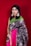 A woman stands against a plain red background, wearing a vibrant saree with intricate patterns, adorned with statement earrings and multiple bangles. The saree is primarily dark blue with metallic details, complemented by a pink border and a green pallu.