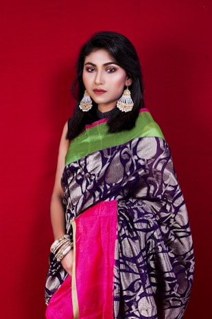 A woman stands against a plain red background, wearing a vibrant saree with intricate patterns, adorned with statement earrings and multiple bangles. The saree is primarily dark blue with metallic details, complemented by a pink border and a green pallu.
