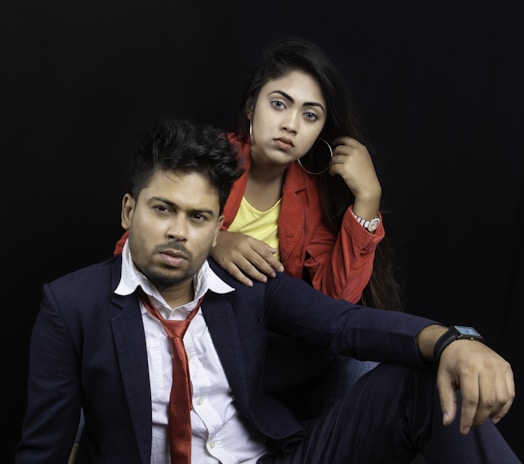 A man and woman are posing against a dark background. The man is wearing a dark suit, a white shirt, and a red tie. He has a serious expression and is sitting with his arm resting on his raised knee. The woman is standing slightly behind him, wearing a red jacket over a yellow top, and is touching her hair. She looks directly at the camera.