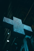 A brightly lit cross sign composed of numerous small LED lights glows in a bluish hue against a dark urban backdrop. The cross is slightly tilted, and there are indistinct structures in the background, hinting at a cityscape.