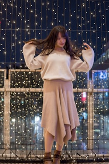 A young woman with long dark hair stands in front of a backdrop of twinkling string lights at night. She wears a cozy, beige sweater and a flowing skirt with a high-low hemline. Her brown ankle boots complement the outfit, and she appears to be playing with her hair while softly smiling.
