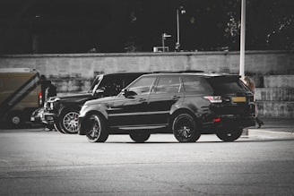 A sleek black SUV with tinted windows is parked on a city street. A similar vehicle is parked nearby, and people are walking in the background. The setting appears urban with a neutral-toned wall and muted surroundings.