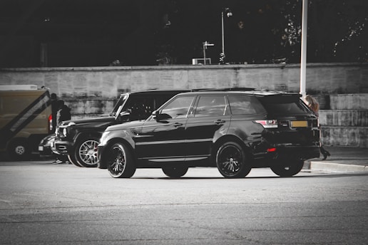 A sleek black SUV with tinted windows is parked on a city street. A similar vehicle is parked nearby, and people are walking in the background. The setting appears urban with a neutral-toned wall and muted surroundings.