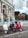 Three scooters are parked side by side in front of a historical fountain. The fountain features ornate arches and appears to be made of stone with Latin inscriptions. The scooters are white, black, and red, and they rest on a cobblestone surface. In the background, there are trees and a cloudy sky.