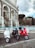Three scooters are parked side by side in front of a historical fountain. The fountain features ornate arches and appears to be made of stone with Latin inscriptions. The scooters are white, black, and red, and they rest on a cobblestone surface. In the background, there are trees and a cloudy sky.