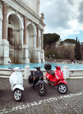 Three scooters are parked side by side in front of a historical fountain. The fountain features ornate arches and appears to be made of stone with Latin inscriptions. The scooters are white, black, and red, and they rest on a cobblestone surface. In the background, there are trees and a cloudy sky.
