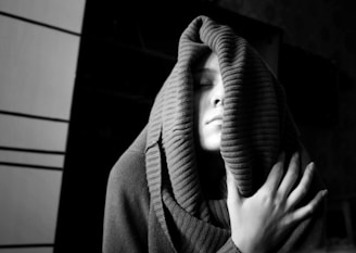 A person with eyes closed, partially covered by a thick, ribbed scarf or fabric, appears to be in a state of calm or warmth. The lighting highlights their face and hand, which rests gently on the scarf, with a mostly dark background giving a sense of intimacy.