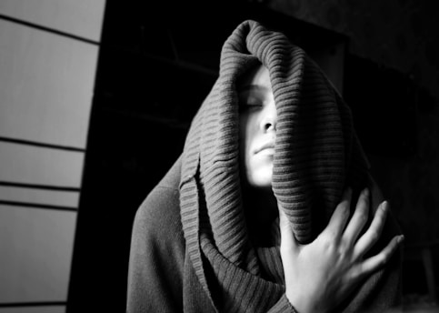A person with eyes closed, partially covered by a thick, ribbed scarf or fabric, appears to be in a state of calm or warmth. The lighting highlights their face and hand, which rests gently on the scarf, with a mostly dark background giving a sense of intimacy.