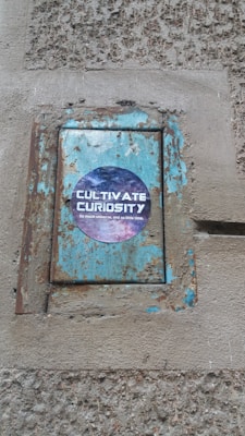 A worn and rusty metal panel on a rough-textured wall with a sticker that reads 'Cultivate Curiosity'. The background of the sticker features a cosmic scene with stars and nebulae.