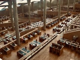The interior of a modern library with rows of wooden desks and computer stations, surrounded by tall, concrete pillars under a high ceiling. There are multiple levels visible, filled with bookshelves and study areas. A few people are seated and working while some are walking along the aisles.