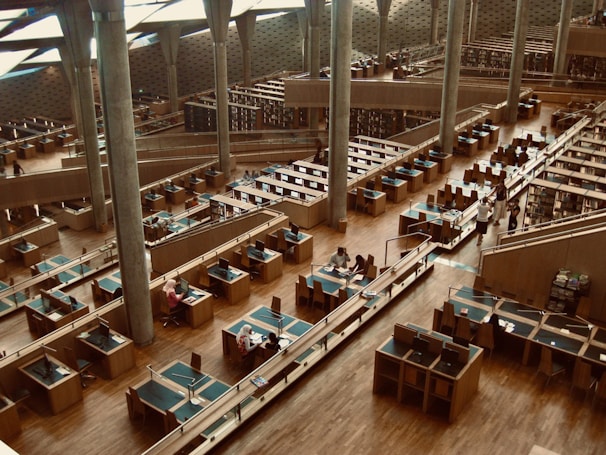 The interior of a modern library with rows of wooden desks and computer stations, surrounded by tall, concrete pillars under a high ceiling. There are multiple levels visible, filled with bookshelves and study areas. A few people are seated and working while some are walking along the aisles.