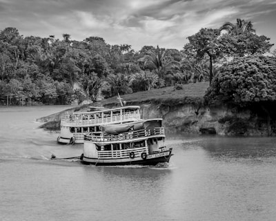 A black and white image featuring a river with two boats traveling through calm waters. Dense, lush trees and foliage line the riverbanks, creating a natural, serene environment. The boats have a classic, vintage design, and one prominently displays the name 'COTIA GILSON III' on its side.