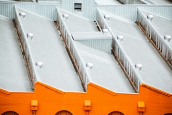A series of industrial roofs with a repeating triangular pattern, featuring a bright orange base with gray metal surfaces. Several small metallic vents are evenly distributed along the rooftops.
