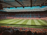A large football stadium filled with spectators showcases an ongoing soccer match. The field is green with well-maintained grass, and players are wearing red and white uniforms. The stands are crowded with fans, and the stadium is equipped with lighting structures above.