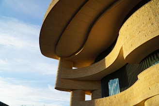 The image features a modern architectural structure with sweeping curves and beige stone textures. The building is illuminated by sunlight, highlighting the intricate design and smooth surfaces. The facade includes vertical glass panels underneath the overhanging sections.