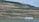 A scenic landscape features terraced vineyards on a hillside, overlooking a river. A white cruise boat sails along the water in the foreground. Lush greenery and a few trees are visible, with a building or monument located at the top of the hill.