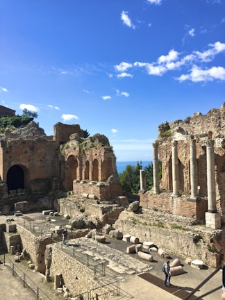 Ancient ruins with large stone columns and brick arches against a bright blue sky. The structure appears to be part of an old theater or temple. A few people are walking along pathways within the ruins surrounded by scattered stone fragments.
