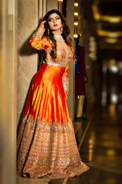 A serene portrait of a woman in elegant terracotta and antique gold attire, standing against a softly lit sandstone wall.