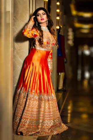 A serene portrait of a woman in elegant terracotta and antique gold attire, standing against a softly lit sandstone wall.