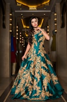 A woman stands elegantly in a luxurious hallway, wearing a long teal gown with intricate gold embroidery. The setting features ornate lighting and mirrors, adding a sophisticated ambiance.