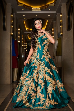 A woman stands elegantly in a luxurious hallway, wearing a long teal gown with intricate gold embroidery. The setting features ornate lighting and mirrors, adding a sophisticated ambiance.