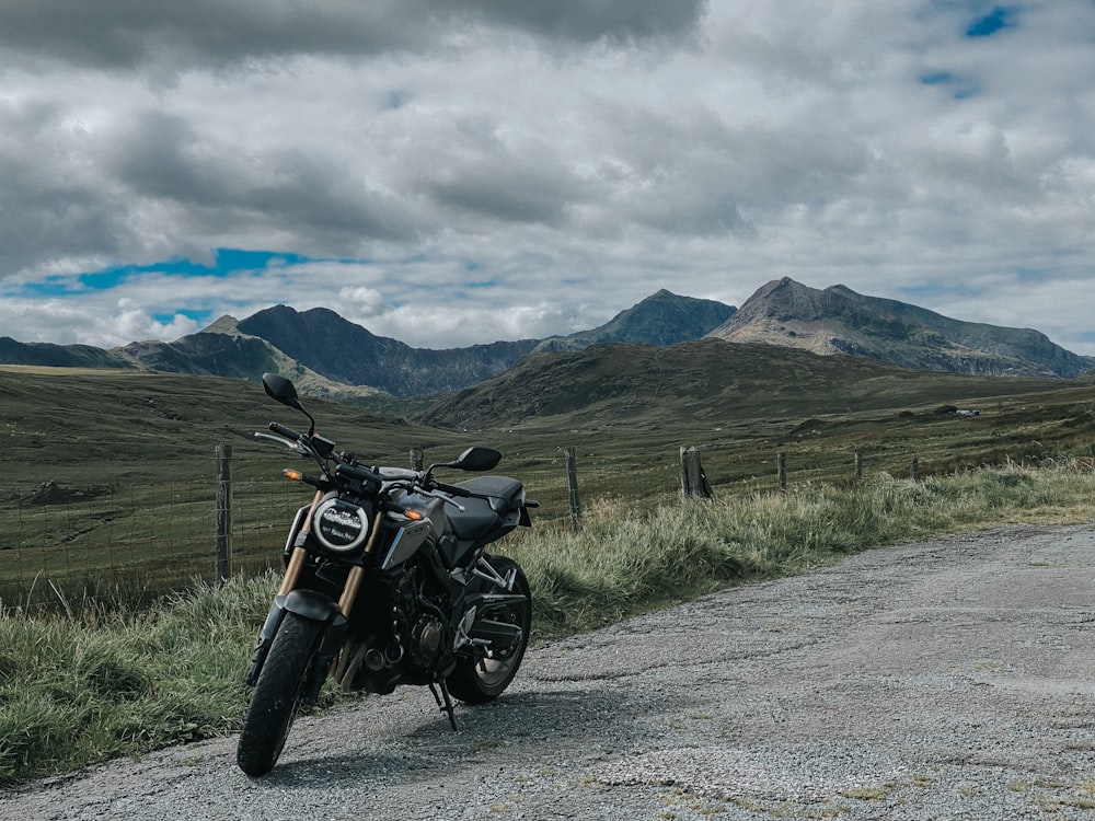 푸른 잔디밭과 흰 구름 아래 산 근처의 회색 아스팔트 도로에 주차된 검은색 오토바이