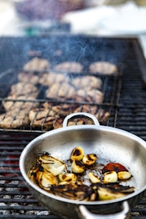 A metal pan filled with grilled and charred vegetables, including onions and garlic, rests on an outdoor barbecue grill. In the background, several pieces of meat are also being cooked over the same grill, with visible smoke rising into the air.