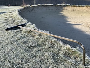 A frost-covered golf course with a large sandy bunker and a metallic rake resting on the frosty grass. In the distance, a person appears to be walking away and carrying a golf bag. The early morning light casts long shadows, adding depth to the scene.